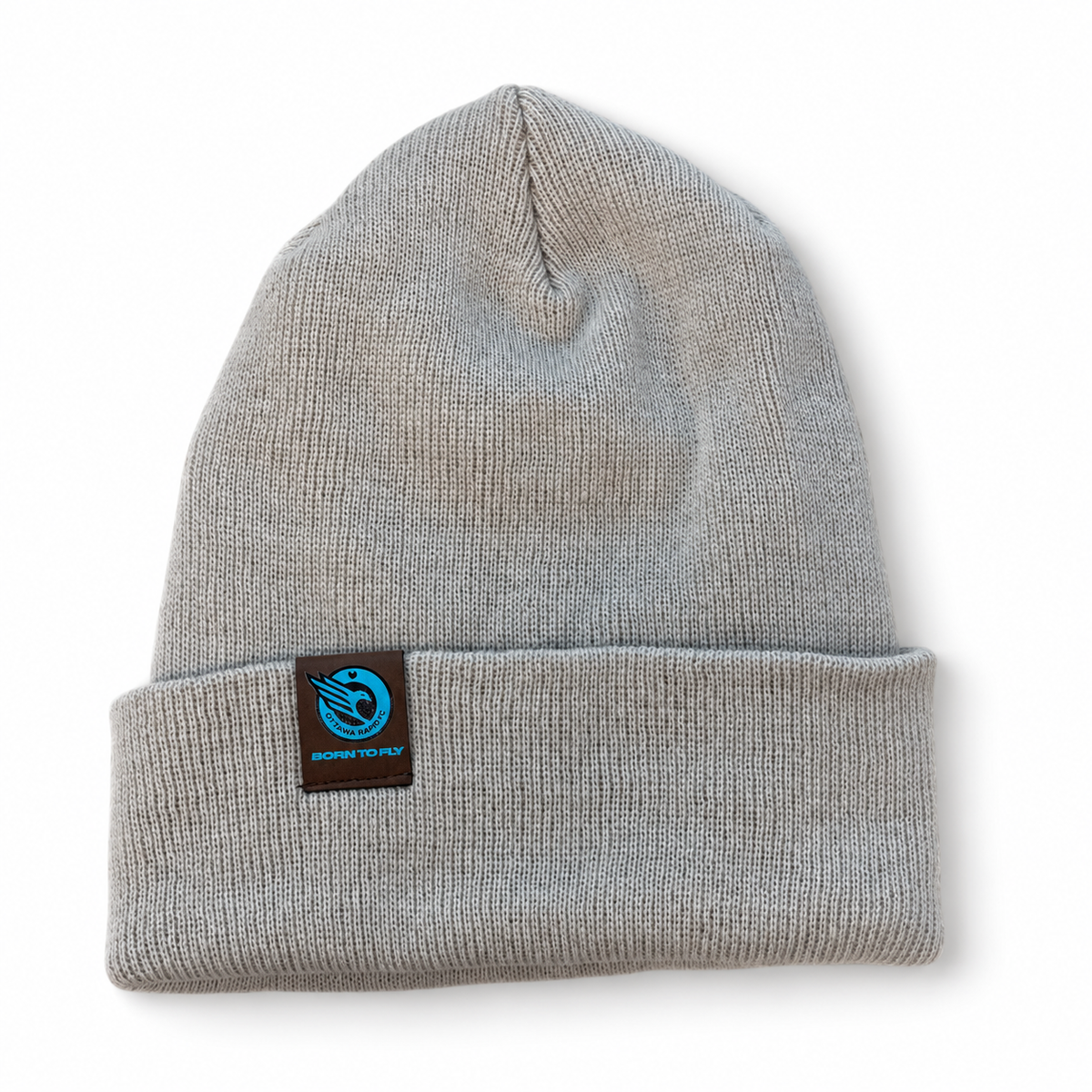 A cream ribbed knit toque with a folded cuff, featuring a small brown leather square patch on the front with the RAPID FC logo and the text 'BORN TO FLY'.